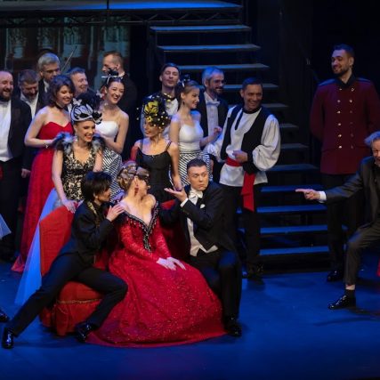 Scena zbiorowa, szeroki kadr. W centrum siedzi kobieta w czerwonej sukni, oczy ma przysłonięte maseczką. Po obu jej stronach siedzą mężczyźni, gestem wskazując na kobietę. Dookoła liczba grupa mężczyzn i kobiet w wystawnych, balowych strojach. Śmieją sie, atmosfera wydaje się pełna zabawy.