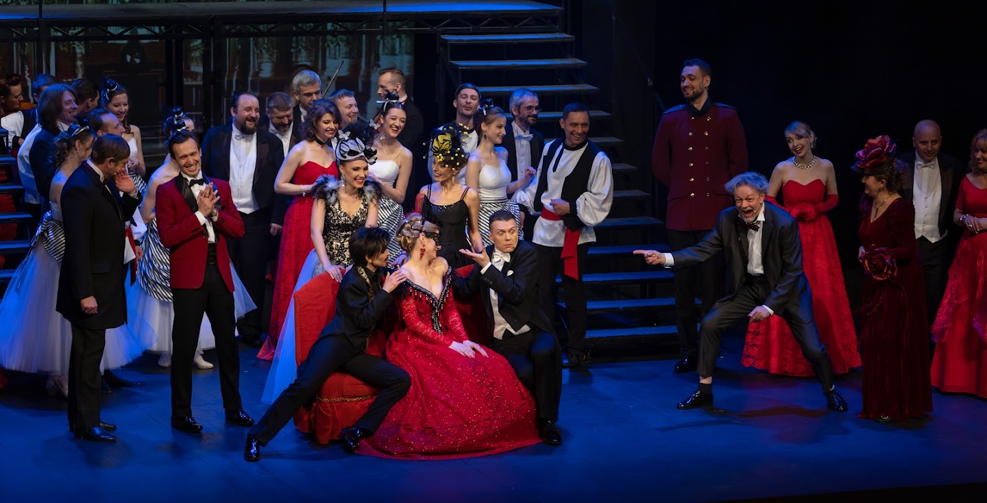 Scena zbiorowa, szeroki kadr. W centrum siedzi kobieta w czerwonej sukni, oczy ma przysłonięte maseczką. Po obu jej stronach siedzą mężczyźni, gestem wskazując na kobietę. Dookoła liczba grupa mężczyzn i kobiet w wystawnych, balowych strojach. Śmieją sie, atmosfera wydaje się pełna zabawy.