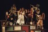 Scena zbiorowa. 8 osób stoi na drewnianych postumentach z ciemnego drewna, przed nimi na podłodze leżą bębny. Osoby są zwrócone w stronę 1 mężczyzny, który klęczy po lewej stronie podestu. Stojący wyciągają dłonie w stronę klęczącego, wydają się śpiewać.