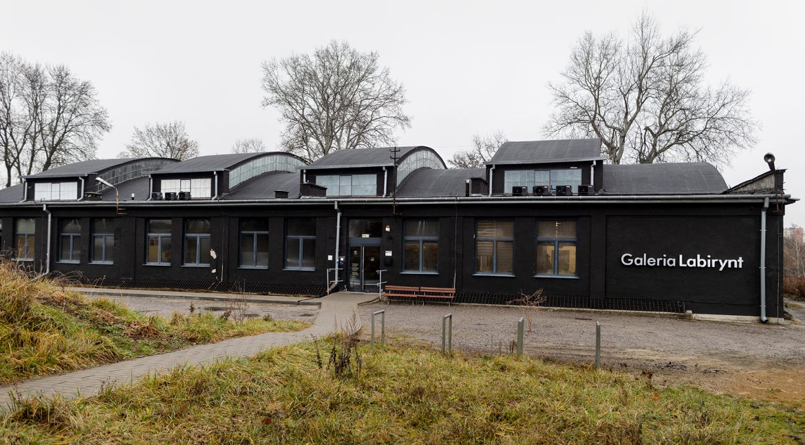 Zdjęcie budynku Galerii Labirynt w Lublinie. Czarna bryła byłego warsztatu samochodowego, na ścianie szczytowej napis białymi literami "Galeria Labirynt".