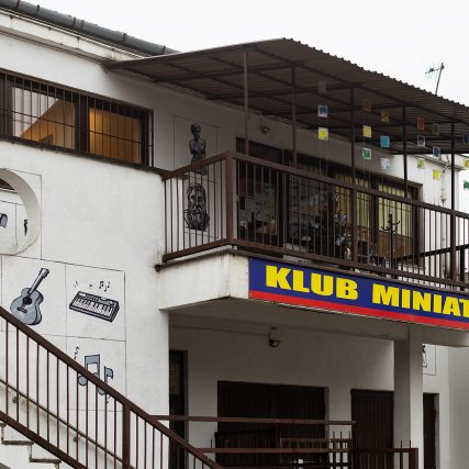 Zdjęcie budynku. Na pierwszym planie balustrada schodów, powyżej balkon przykryty blachą falistą. Pod balustradą balkonu napis "Klub Minatura".